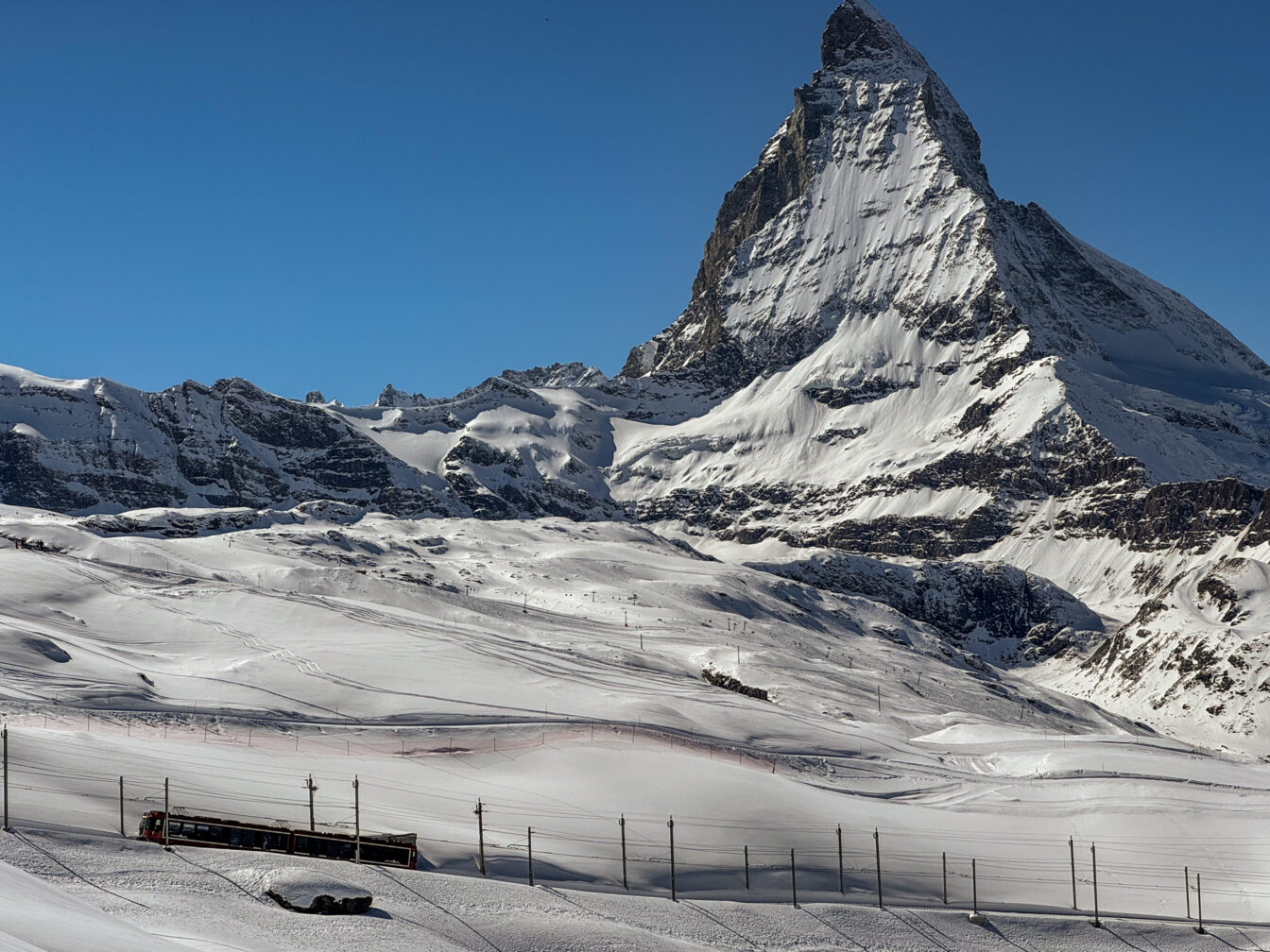 Winterlandschaft in Zermatt mit dem Matterhorn im Hintergrund. Im Vordergrund fährt eine rote Zahnradbahn durch das schneebedeckte Skigebiet, während Skipisten und Spuren im Schnee sichtbar sind. Strahlend blauer Himmel ohne Wolken.