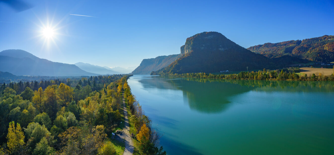 Sonniger Herbsttag am Drauradweg in Kärnten: Ein breiter Fluss schlängelt sich durch das Tal, flankiert von buntem Laubwald und einem Radweg. Im Hintergrund erheben sich bewaldete Berge unter blauem Himmel
