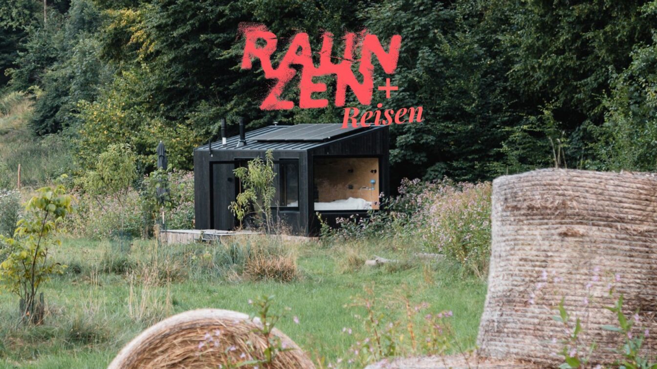 Ein kleines schwarzes Holzhaus mit großen Fenstern steht inmitten einer grünen Wiese am Waldrand. Im Vordergrund liegen große Heuballen, im Hintergrund dichter Wald. Auf dem Bild ist in roter Schrift der Schriftzug 'RAUM ZEN + Reisen' zu sehen