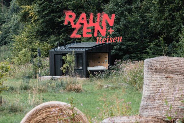 Ein kleines schwarzes Holzhaus mit großen Fenstern steht inmitten einer grünen Wiese am Waldrand. Im Vordergrund liegen große Heuballen, im Hintergrund dichter Wald. Auf dem Bild ist in roter Schrift der Schriftzug 'RAUM ZEN + Reisen' zu sehen