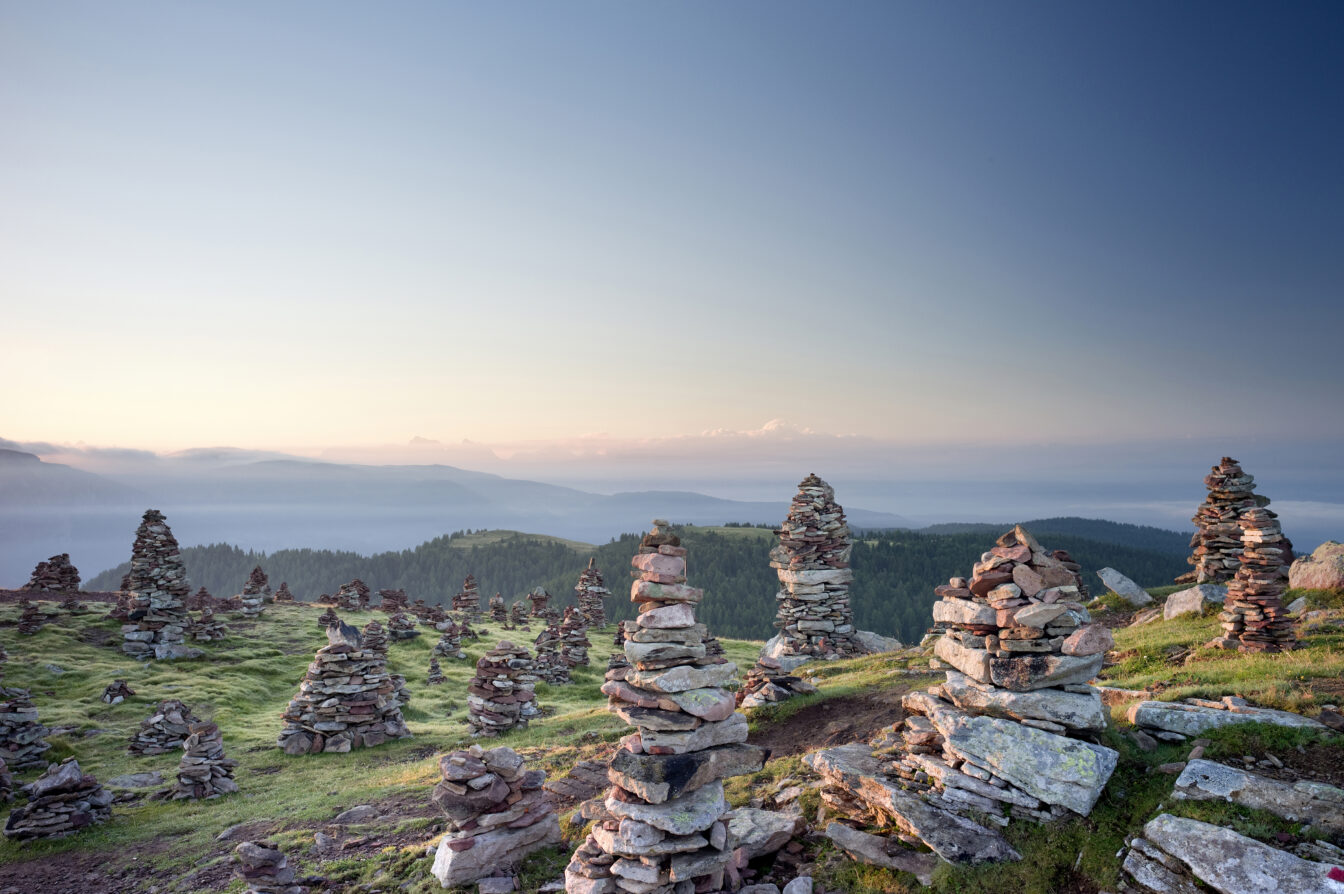 Zahlreiche aufgestapelte Steintürme im Sarntal, die in der Abendsonne leuchten, mit Blick auf bewaldete Hügel und ferne Bergketten unter einem klaren Himmel