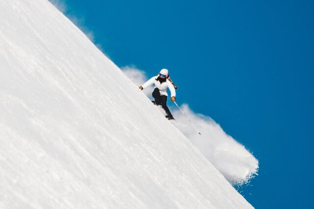 Skifahren in Bayern auf einer steilen Piste bei strahlend blauem Himmel – perfekte Wintersportbedingungen in beliebten Skigebieten wie Garmisch-Partenkirchen, Spitzingsee oder Sudelfeld, die bequem mit den Öffentlichen Verkehrsmitteln erreichbar sind.