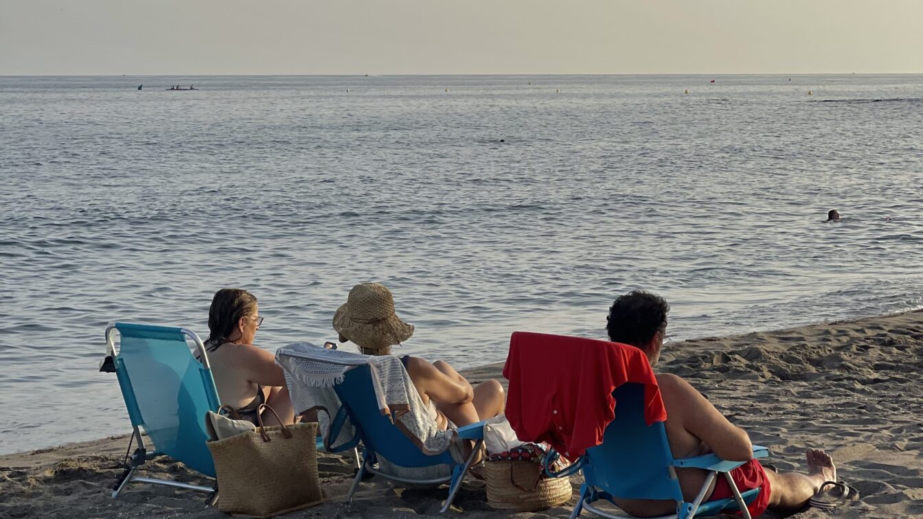 Drei Menschen sitzen auf Liegestühlen am Strand. Es ist Abendstimmung.