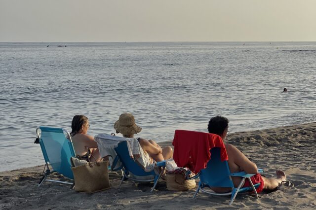 Drei Menschen sitzen auf Liegestühlen am Strand. Es ist Abendstimmung.