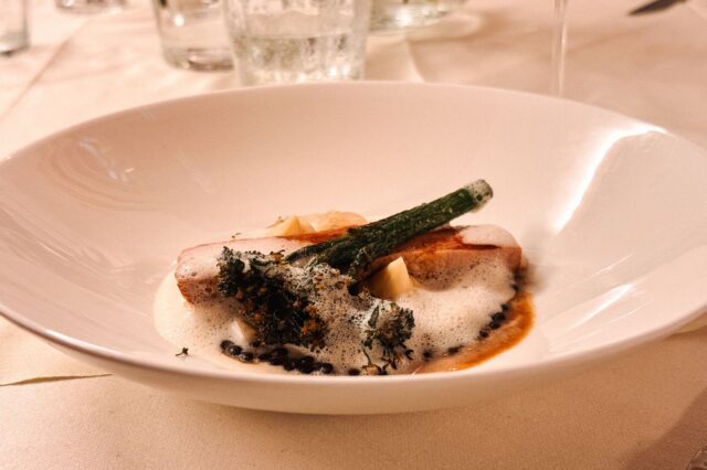 Gourmetgericht im Restaurant Triad in der Buckligen Welt: Zart gebratenes Fleisch mit Brokkolini, cremigem Schaum, Linsen und feiner Sauce, kunstvoll angerichtet auf weißem Porzellanteller.