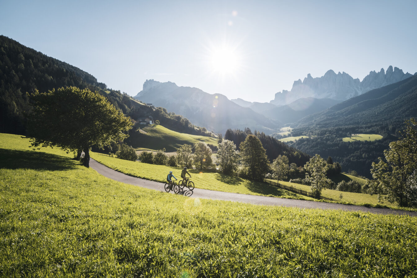 Zwei Radfahrer auf einem sonnigen Weg durch grüne Wiesen im Villnößtal, umgeben von Wäldern und den markanten Dolomitengipfeln im Hintergrund.