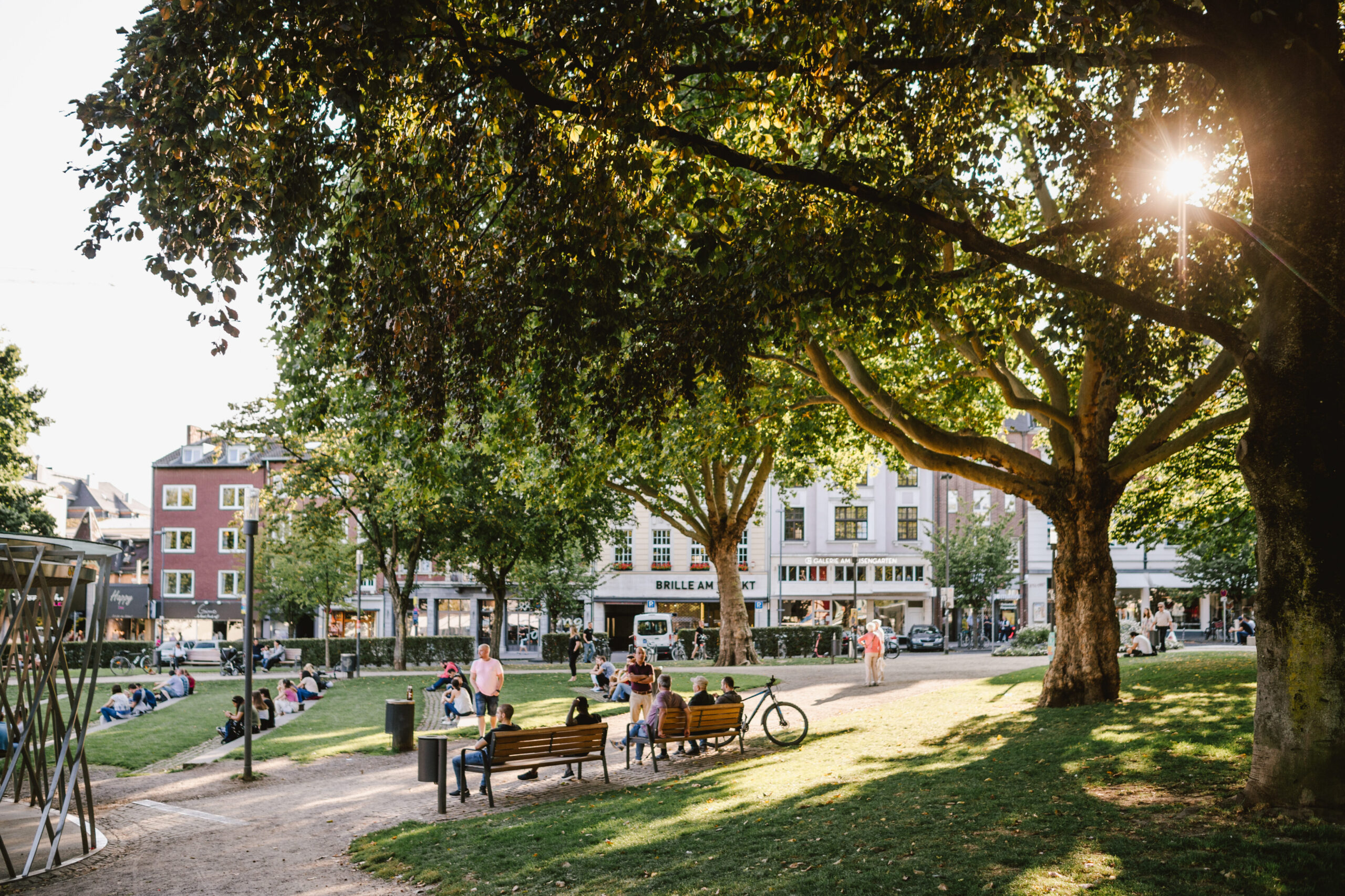 Menschen genießen den sonnigen Nachmittag im Elisengarten in Aachen, sitzen auf Bänken und im Gras unter großen, schattenspendenden Bäumen mit Blick auf umliegende Altstadthäuser.