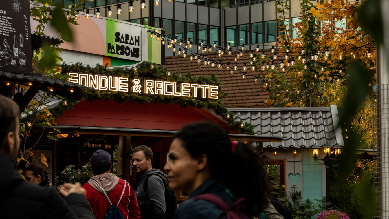 Belebte Adventsszene in Zürich mit einem festlich geschmückten Stand, auf dem in leuchtenden Buchstaben ‚Fondue & Raclette‘ steht. Menschen schlendern davor entlang, während über ihnen Lichterketten hängen. Im Hintergrund sind herbstlich gefärbte Bäume und moderne Gebäude zu sehen.