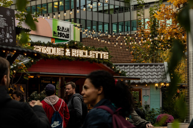 Belebte Adventsszene in Zürich mit einem festlich geschmückten Stand, auf dem in leuchtenden Buchstaben ‚Fondue & Raclette‘ steht. Menschen schlendern davor entlang, während über ihnen Lichterketten hängen. Im Hintergrund sind herbstlich gefärbte Bäume und moderne Gebäude zu sehen.