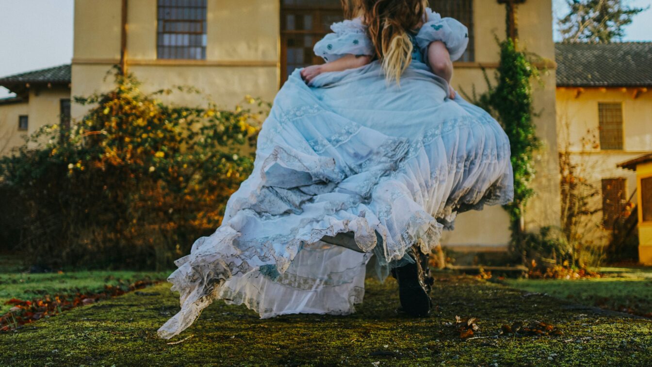 Eine Person in einem langen, hellblauen, verspielten Kleid läuft oder tanzt über einen moosbewachsenen Weg vor einem alten Gebäude. Der Stil erinnert an ein Märchen oder eine Szene aus Alice im Wunderland.