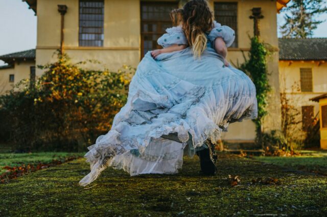 Eine Person in einem langen, hellblauen, verspielten Kleid läuft oder tanzt über einen moosbewachsenen Weg vor einem alten Gebäude. Der Stil erinnert an ein Märchen oder eine Szene aus Alice im Wunderland.