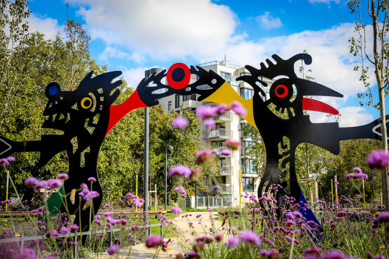 Farbenfrohe Skulpturen von André Heller im Kunstpark in Wien, eingerahmt von blühenden Blumen und moderner Architektur im Hintergrund.