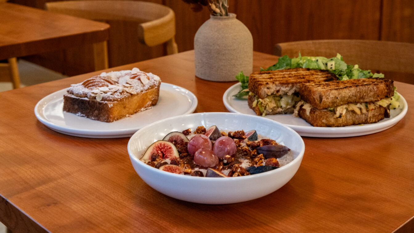 Ein gemütlich gedeckter Holztisch in einem Café in Zürich mit drei Brunchgerichten: ein Teller mit einem Stück Mandelkuchen, ein Teller mit einem gegrillten Sandwich und Salat, sowie eine Schüssel mit Porridge, frischen Feigen, Trauben und Granola. Im Hintergrund stehen Holzstühle und eine Vase mit Trockenblumen.