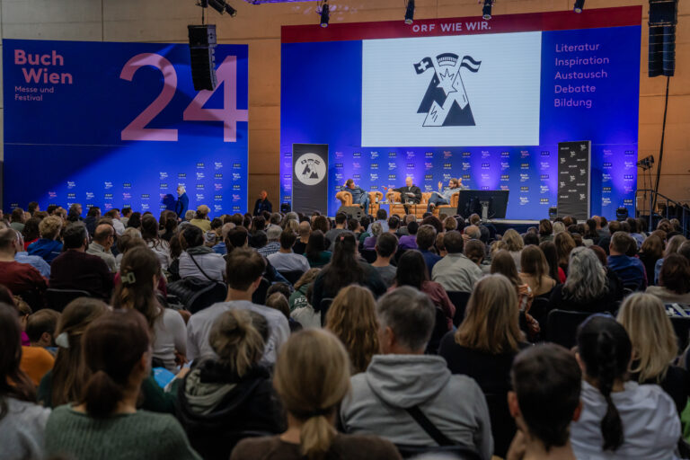 Publikum bei einer Podiumsdiskussion auf der Buch Wien 2024. Auf der Bühne sitzen mehrere Personen in Sesseln, hinter ihnen eine große Leinwand mit Logo und Schriftzug. Links und rechts sind große blaue Banner mit der Aufschrift ‚Buch Wien – Messe und Festival‘ sowie ‚Literatur, Inspiration, Austausch, Debatte, Bildung‘