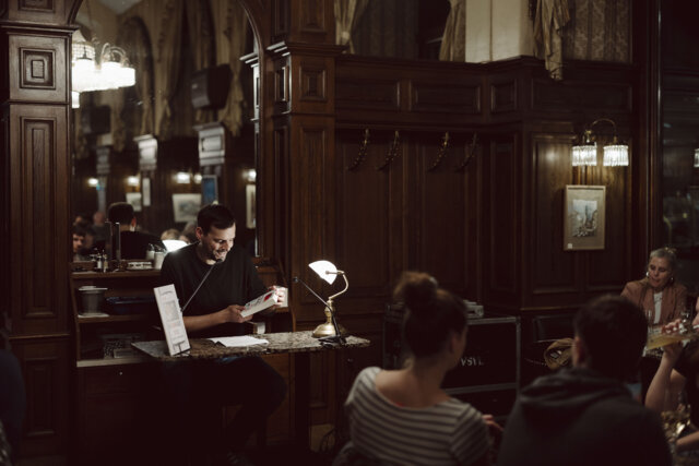 Ein Mann liest bei der Kriminacht in Wien in einem holzvertäfelten Raum eines Kaffeehauses aus einem Buch, während mehrere Personen im Publikum aufmerksam zuhören.