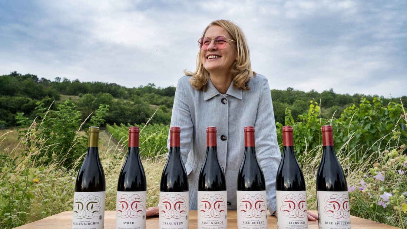 Eine lachende Frau mit blonden Haaren und rosa Sonnenbrille steht hinter einem Holztisch mit sieben Weinflaschen. Jede Flasche trägt ein Etikett mit dem Namen des Weinguts Dorli Muhr und unterschiedlichen Sorten. Im Hintergrund sind grüne Weinberge unter einem bewölkten Himmel zu sehen.