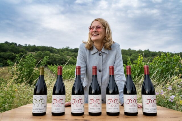 Eine lachende Frau mit blonden Haaren und rosa Sonnenbrille steht hinter einem Holztisch mit sieben Weinflaschen. Jede Flasche trägt ein Etikett mit dem Namen des Weinguts Dorli Muhr und unterschiedlichen Sorten. Im Hintergrund sind grüne Weinberge unter einem bewölkten Himmel zu sehen.
