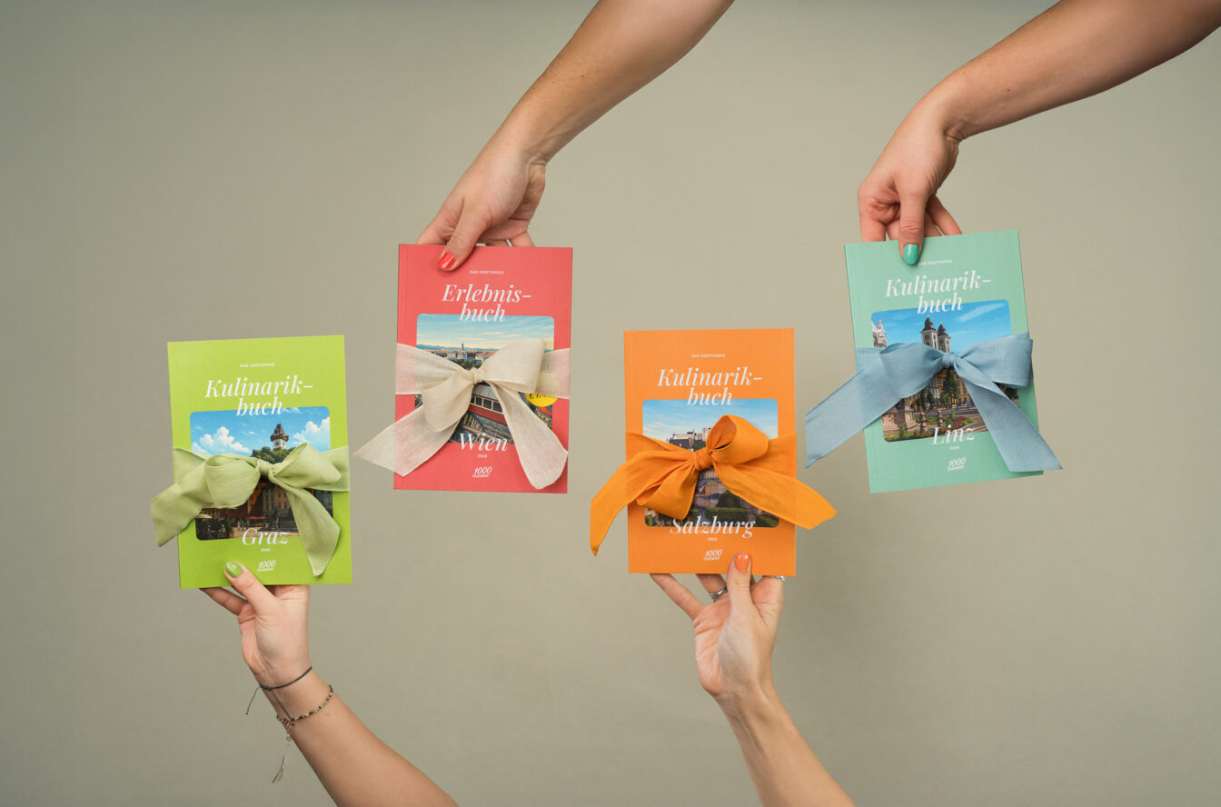 Vier Hände halten die 1000things Bücher – Erlebnisbuch Wien und Kulinarikbücher Graz, Salzburg und Linz – jeweils mit farbigen Geschenk-Schleifen.