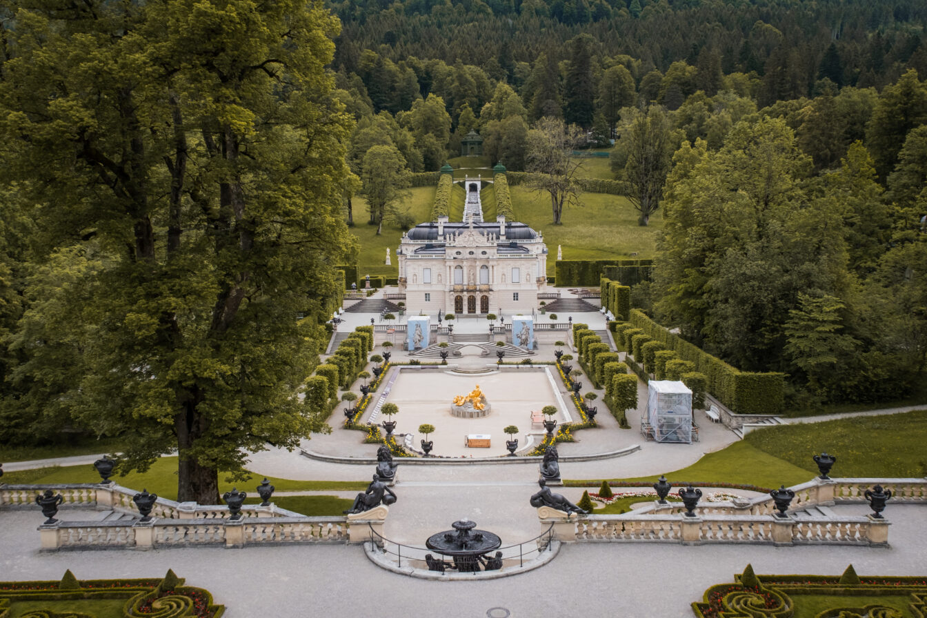 Luftaufnahme von Schloss Linderhof bei Ettal in Bayern, umgeben von symmetrischen Gartenanlagen, Springbrunnen und dichtem Wald im Hintergrund.