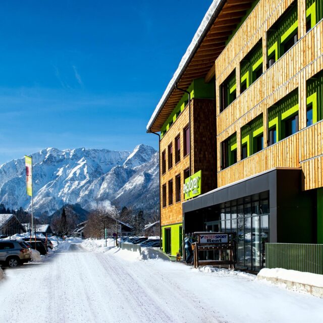 Auf der rechten Seite ist ein Hotel zu sehen, es liegt Schnee und im Hintergrund liegt ein Gebirge.
