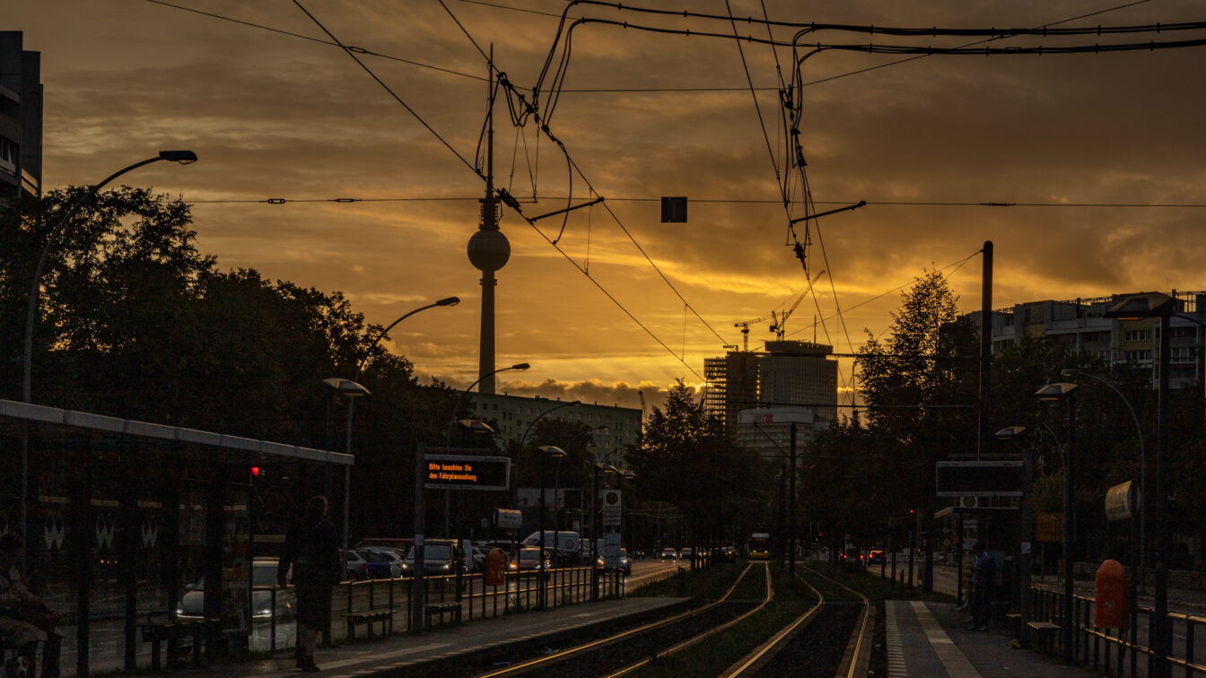 Straßenbahnhaltestelle in Berlin bei Sonnenuntergang, mit Gleisen im Vordergrund, Fahrgästen an den Bahnsteigen, Oberleitungen und der Berliner Fernsehturm im Hintergrund. Ein elektronisches Anzeigeschild zeigt Fahrplaninformationen.