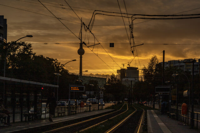 Straßenbahnhaltestelle in Berlin bei Sonnenuntergang, mit Gleisen im Vordergrund, Fahrgästen an den Bahnsteigen, Oberleitungen und der Berliner Fernsehturm im Hintergrund. Ein elektronisches Anzeigeschild zeigt Fahrplaninformationen.