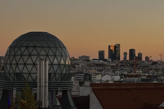 Städtische Skyline von Wien bei Sonnenuntergang – im Vordergrund eine Kuppel mit Glasstruktur und roten Ziegeldächern, im Hintergrund moderne Hochhäuser unter einem orange-blauen Himmel