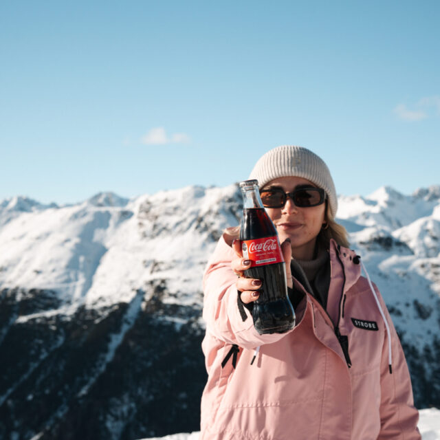 Person hält eine Flasche Cola am schneebedeckten Berg im Winteroutfit