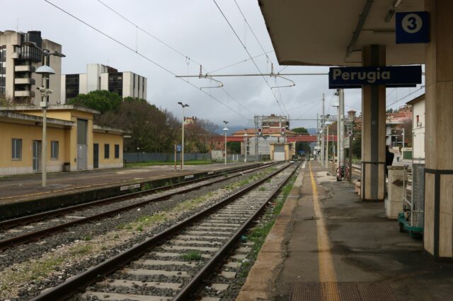 Bahngleise und Bahnsteig am Bahnhof Perugia in Italien, mit Gebäuden im Hintergrund und bewölktem Himmel.