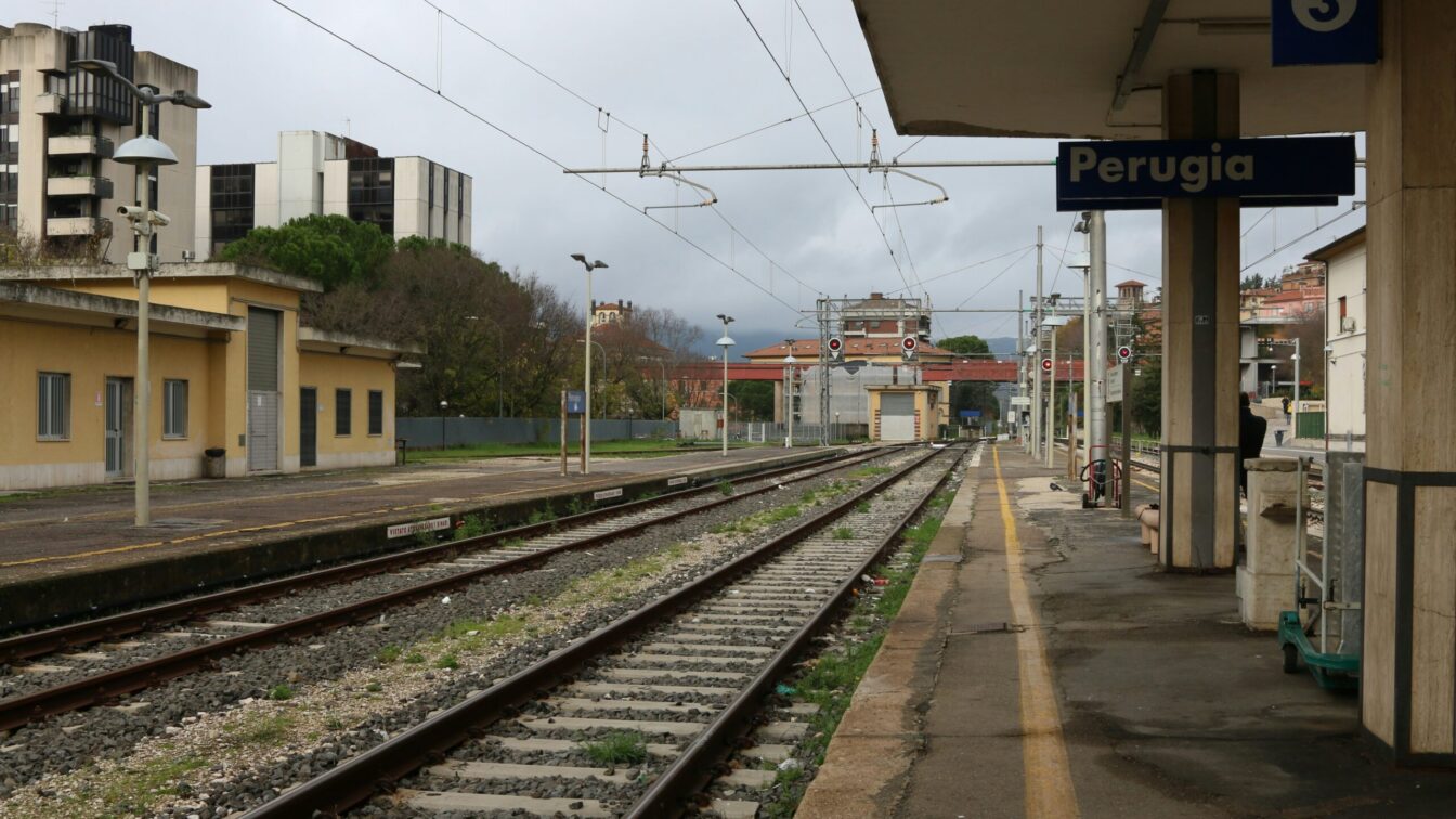 Bahngleise und Bahnsteig am Bahnhof Perugia in Italien, mit Gebäuden im Hintergrund und bewölktem Himmel.