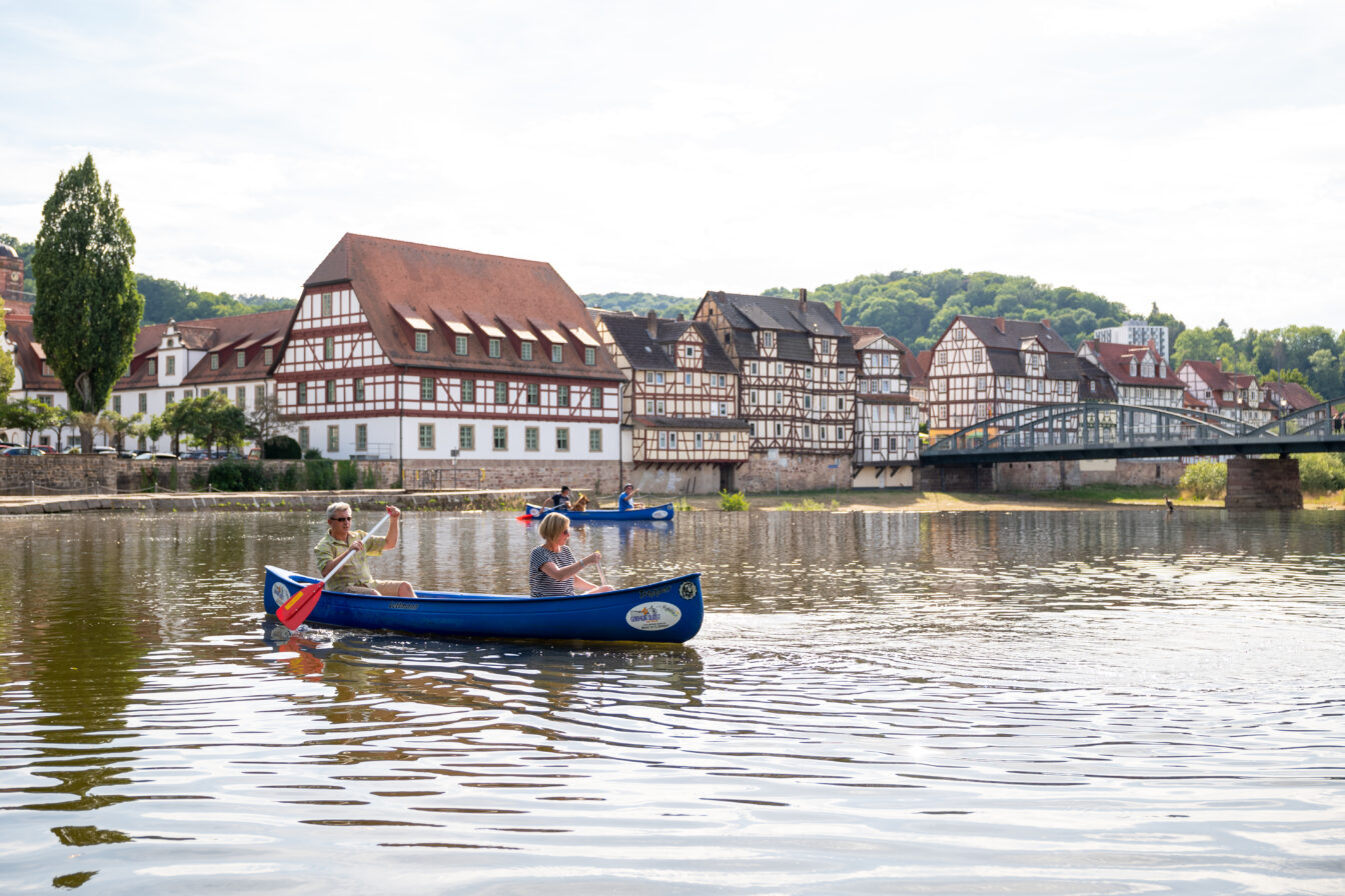 Zwei Personen paddeln in einem blauen Kanu auf einem Fluss, im Hintergrund Fachwerkhäuser und eine Brücke in einer historischen Altstadt bei sonnigem Wetter.
