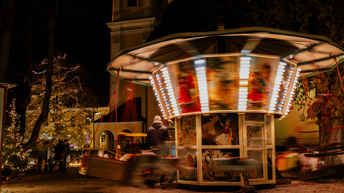 Nostalgisches Kinderkarussell mit bunten Lichtern dreht sich bei Nacht auf dem Adventmarkt in Mondsee, umgeben von festlich beleuchteten Bäumen und weihnachtlicher Atmosphäre.