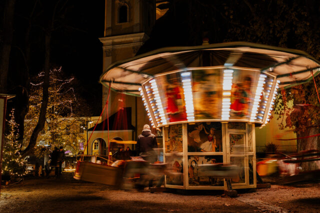 Nostalgisches Kinderkarussell mit bunten Lichtern dreht sich bei Nacht auf dem Adventmarkt in Mondsee, umgeben von festlich beleuchteten Bäumen und weihnachtlicher Atmosphäre.