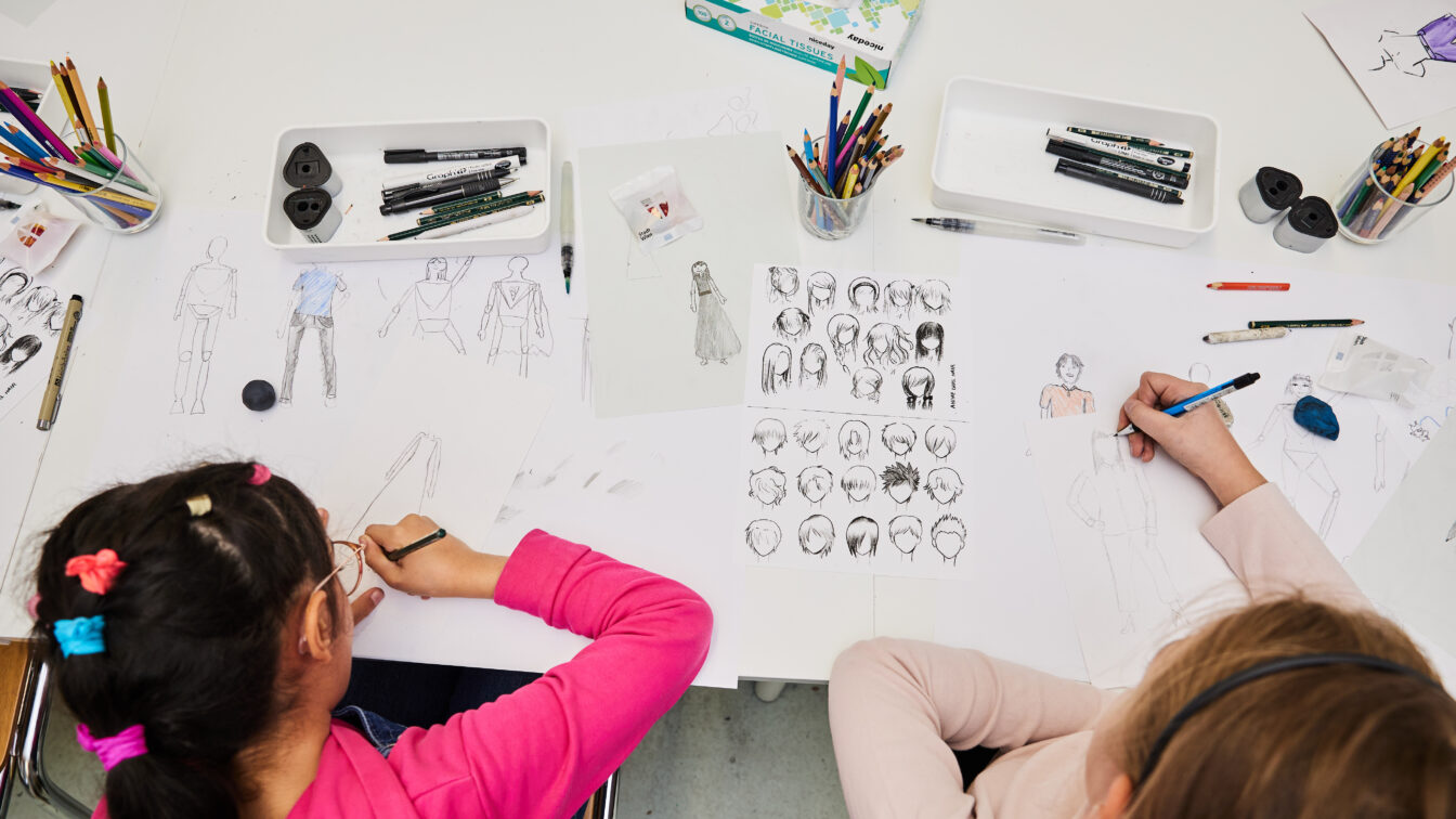 Zwei Kinder sitzen an einem Tisch und zeichnen Figuren und Porträts auf Papier. Auf dem Tisch liegen viele Bunt- und Bleistifte, Fineliner, Radiergummis und Skizzenblätter mit verschiedenen Körperhaltungen und Frisuren. Die Szene wirkt kreativ und konzentriert – ein Moment aus einem Kinderkunstfest oder Zeichenworkshop.
