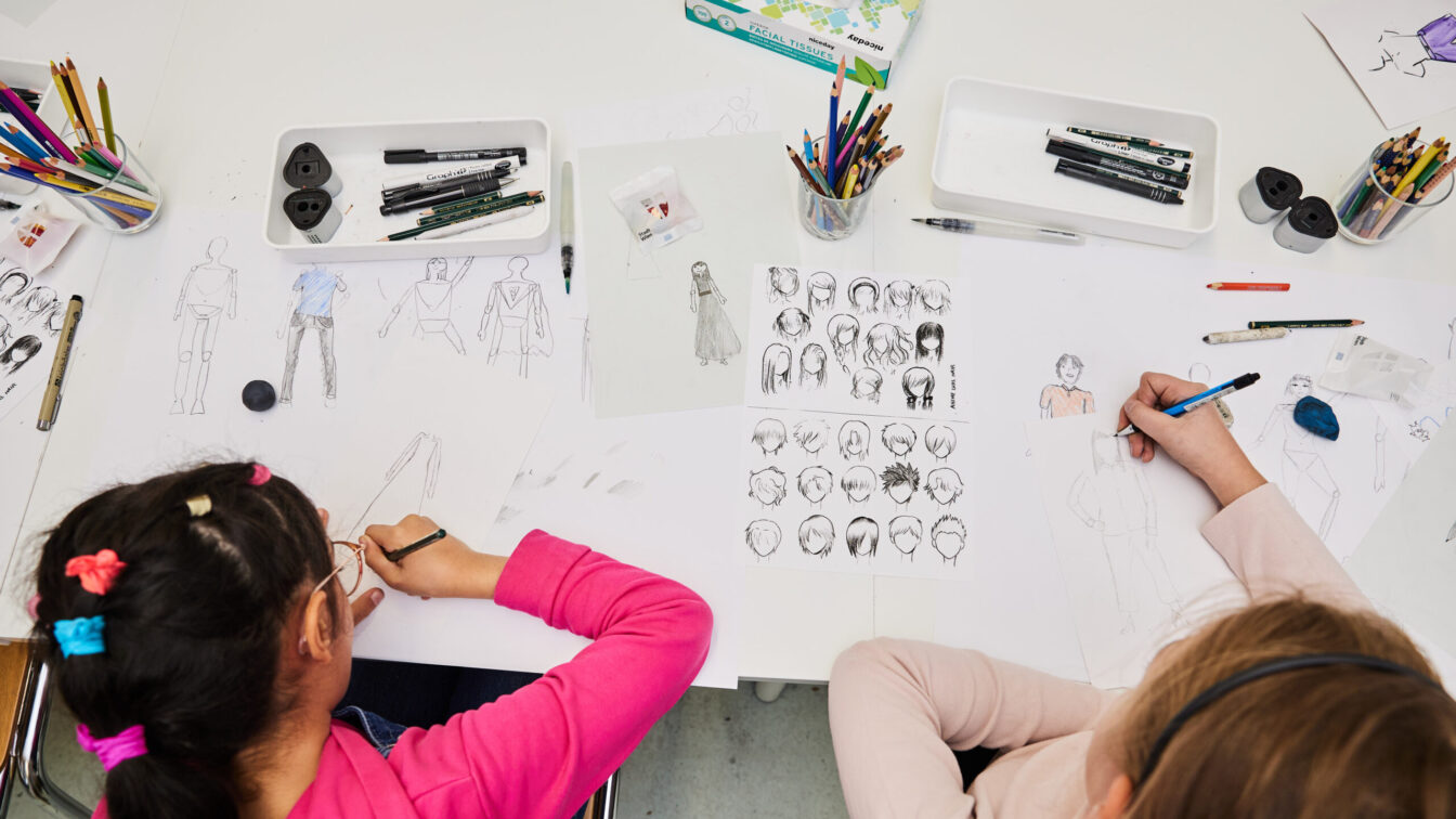 Zwei Kinder sitzen an einem Tisch und zeichnen Figuren und Porträts auf Papier. Auf dem Tisch liegen viele Bunt- und Bleistifte, Fineliner, Radiergummis und Skizzenblätter mit verschiedenen Körperhaltungen und Frisuren. Die Szene wirkt kreativ und konzentriert – ein Moment aus einem Kinderkunstfest oder Zeichenworkshop.