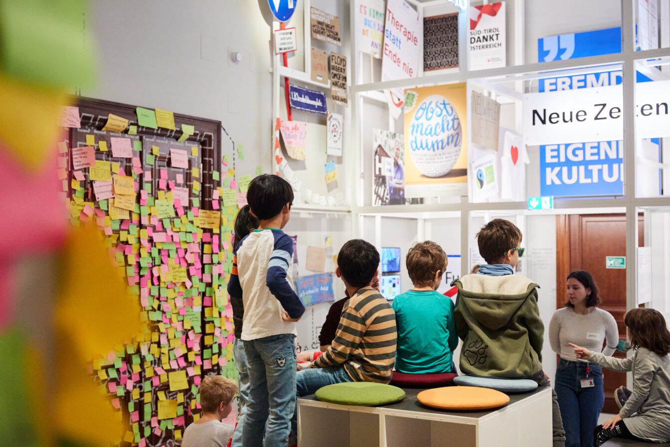 Mehrere Kinder sitzen und stehen in einem hellen Raum voller bunter Plakate, Poster und Botschaften an den Wänden. Eine Wand ist mit vielen farbigen Post-its bedeckt, auf denen Wörter und Ideen stehen. Eine Erwachsene spricht mit der Gruppe – die Szene zeigt ein kreatives und interaktives Kinderkunstfest oder Workshop, bei dem Kinder ihre Gedanken und Wünsche ausdrücken.