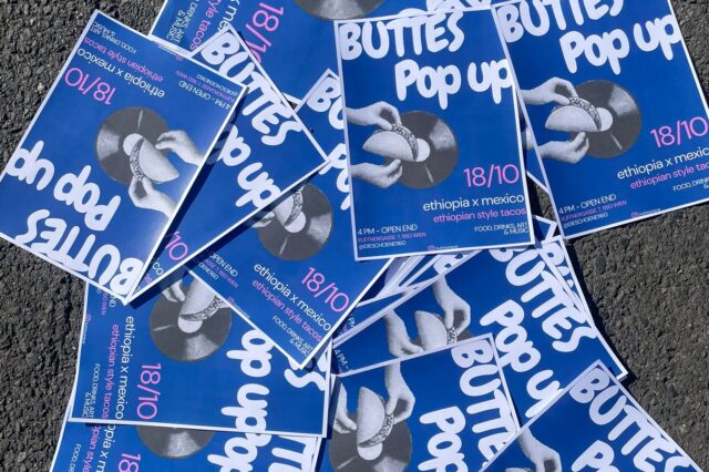 Mehrere blaue Plakate liegen auf Asphalt verteilt. Auf den Plakaten steht in weißer Schrift ‚BUTTES Pop up‘ mit dem Datum 18/10. Darunter steht ‚ethiopia x mexico – ethiopian style tacos‘ sowie weitere Veranstaltungsinformationen. In der Mitte ist ein Schwarz-Weiß-Foto von zwei Händen, die einen Taco halten