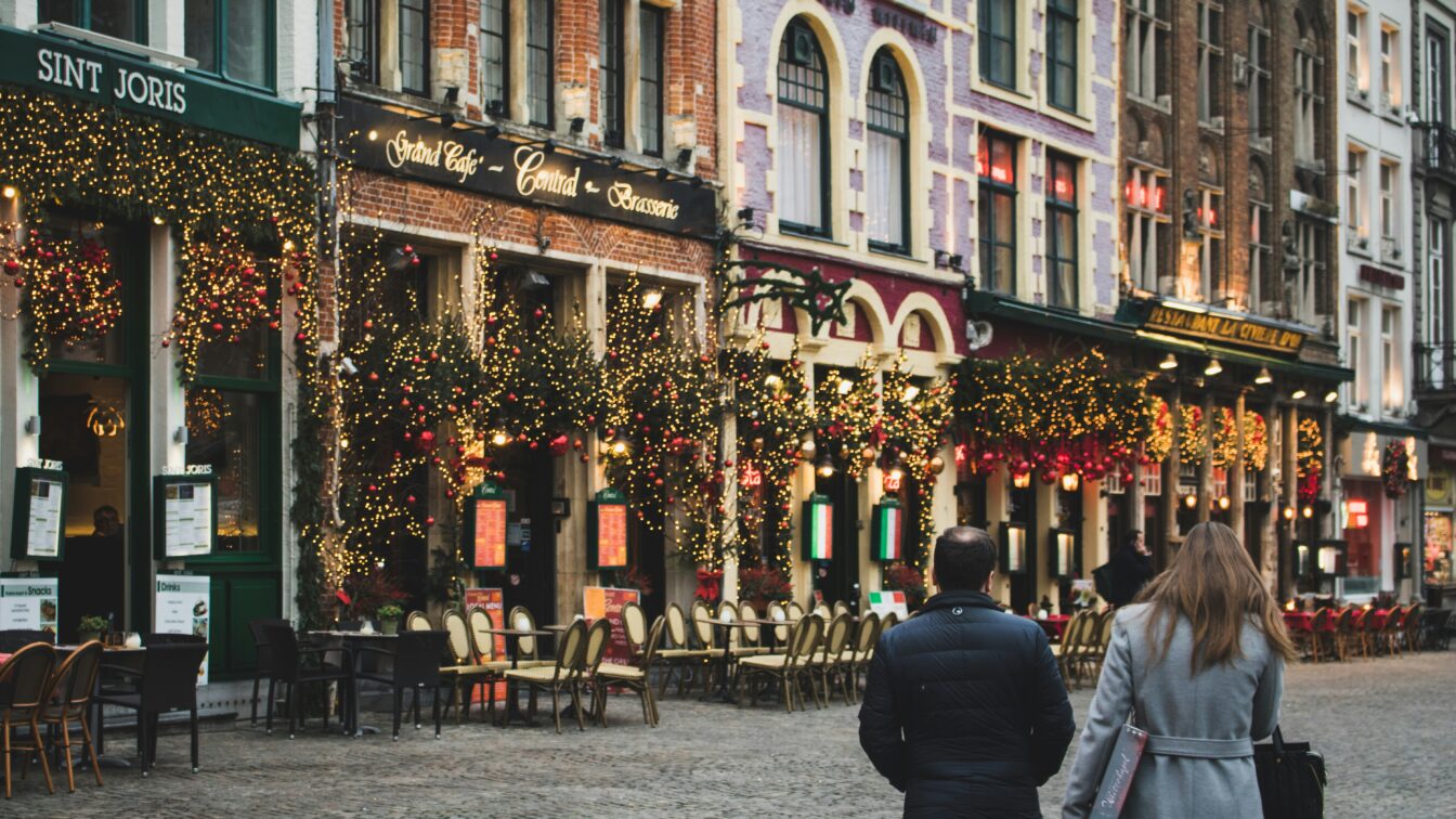 Stimmungsvolle europäische Altstadt im Dezember mit festlich geschmückten Gebäuden, Lichterketten und Weihnachtsdekorationen vor gemütlichen Cafés und Restaurants; zwei Menschen spazieren über das Kopfsteinpflaster – winterliche, weihnachtliche Atmosphäre.