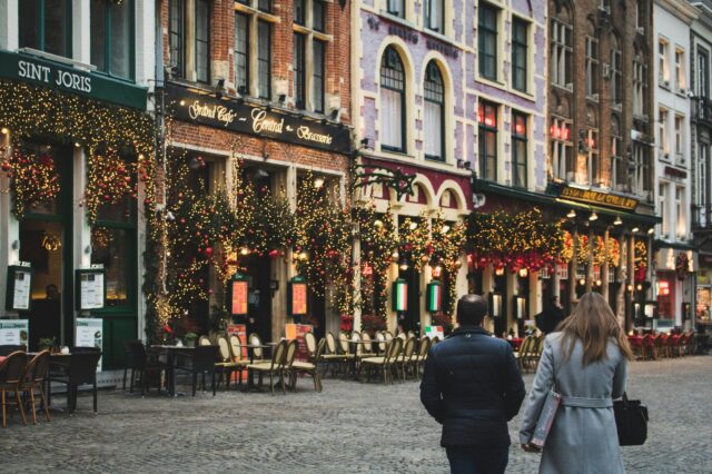 Stimmungsvolle europäische Altstadt im Dezember mit festlich geschmückten Gebäuden, Lichterketten und Weihnachtsdekorationen vor gemütlichen Cafés und Restaurants; zwei Menschen spazieren über das Kopfsteinpflaster – winterliche, weihnachtliche Atmosphäre.