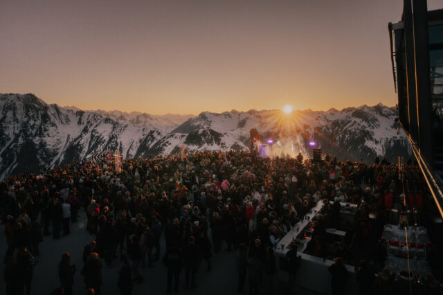 Eine große Menschenmenge feiert bei Sonnenuntergang auf einer Bergplattform umgeben von schneebedeckten Alpen. Im Vordergrund steht eine Bar, im Hintergrund eine Bühne mit Lichtshow. Das Bild zeigt die festliche Atmosphäre eines Ski-Openings 2025 in einem österreichischen Skigebiet