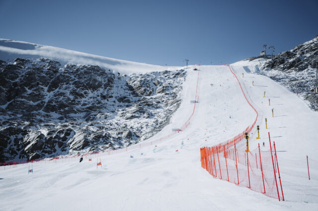 Ein schneebedeckter Hang im Skigebiet Sölden mit roter Sicherheitsabsperrung und Slalomtoren für ein Weltcuprennen. Mehrere Skifahrer sind auf der präparierten Piste unterwegs, umgeben von felsigen Berghängen unter blauem Himmel.