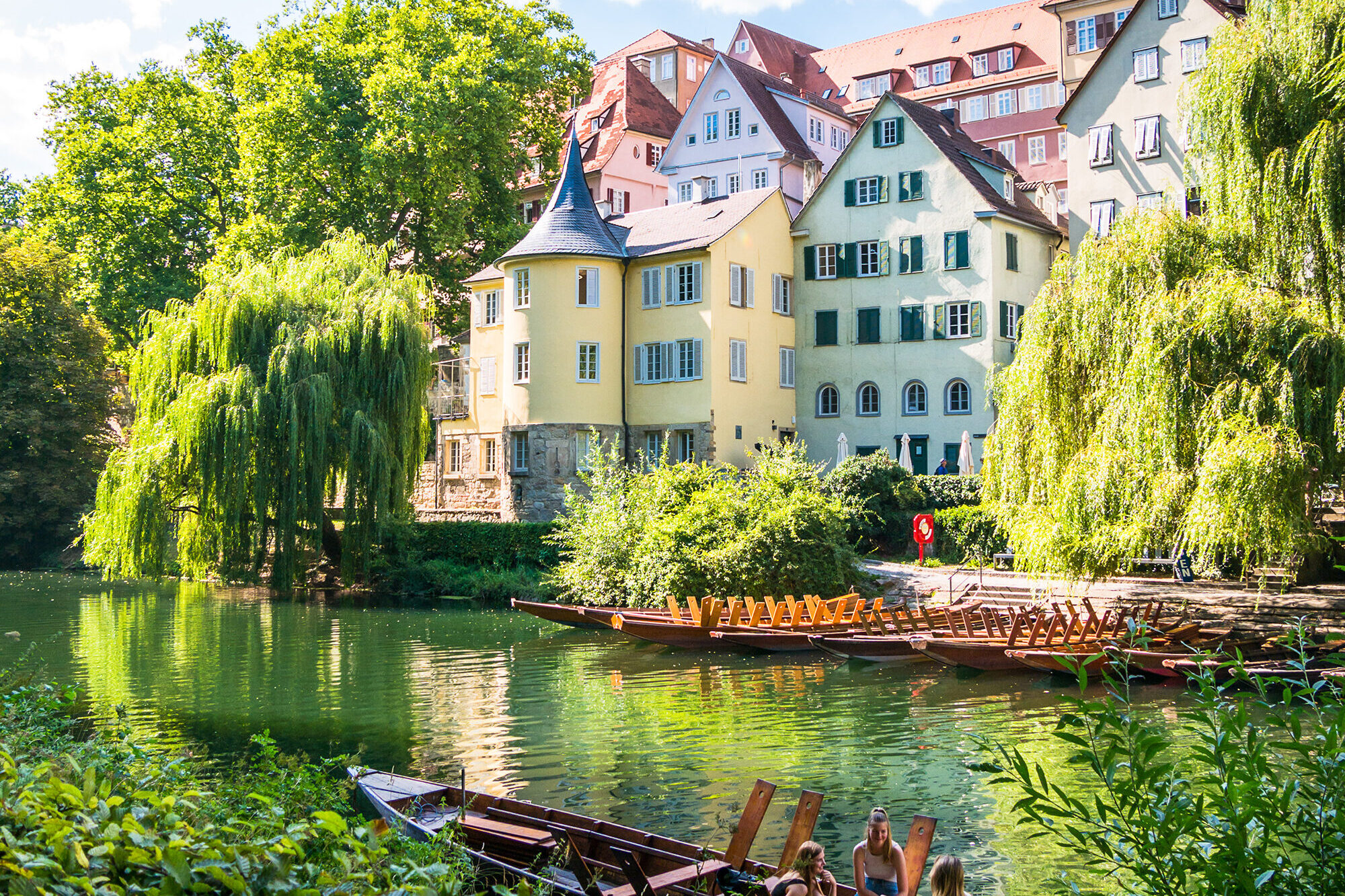 Blick auf den Hölderlinturm und bunte Altstadthäuser am Neckarufer in Tübingen, umgeben von üppigem Grün; im Vordergrund sitzen Menschen in einem Stocherkahn auf dem Wasser bei sonnigem Wetter.