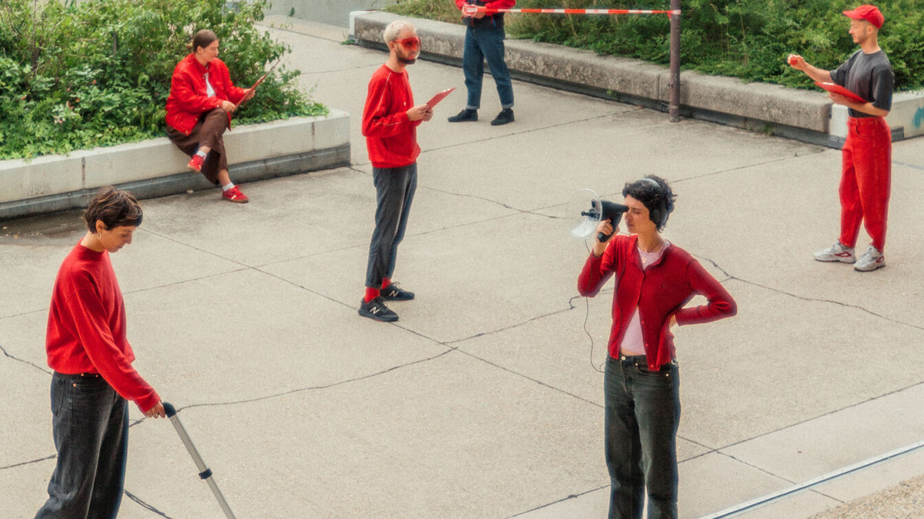 Mehrere Personen in roter Kleidung stehen und sitzen auf einer gepflasterten Freifläche im Freien, umgeben von grünen Pflanzen. Eine Person misst den Boden mit einem Messrad, eine andere spricht durch ein Megafon. Andere halten Klemmbretter oder notieren etwas. Die Szene wirkt wie eine performative Aktion oder eine partizipative Stadtforschungsaktivität im öffentlichen Raum.