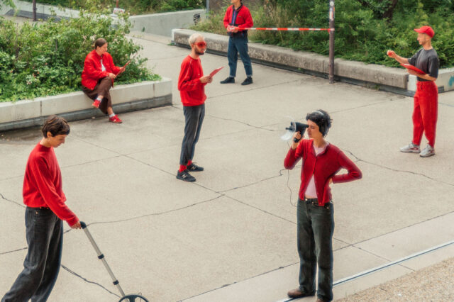 Mehrere Personen in roter Kleidung stehen und sitzen auf einer gepflasterten Freifläche im Freien, umgeben von grünen Pflanzen. Eine Person misst den Boden mit einem Messrad, eine andere spricht durch ein Megafon. Andere halten Klemmbretter oder notieren etwas. Die Szene wirkt wie eine performative Aktion oder eine partizipative Stadtforschungsaktivität im öffentlichen Raum.