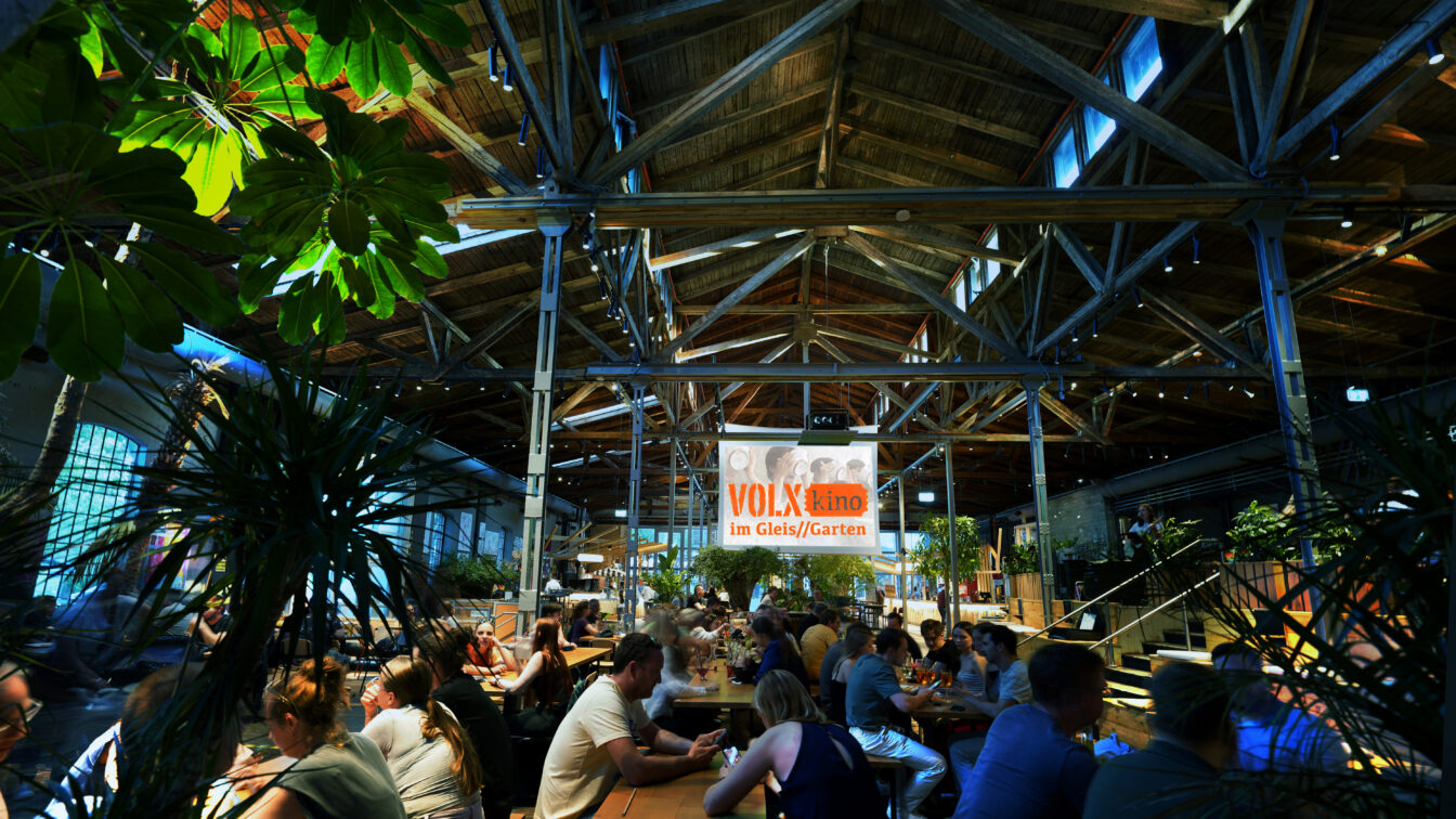 Innenaufnahme des VOLXkino im Gleis/Garten 2025: Menschen sitzen an langen Holztischen in einer großen, offenen Halle mit Holzdachkonstruktion, viel Grünpflanzen und gemütlicher Atmosphäre.