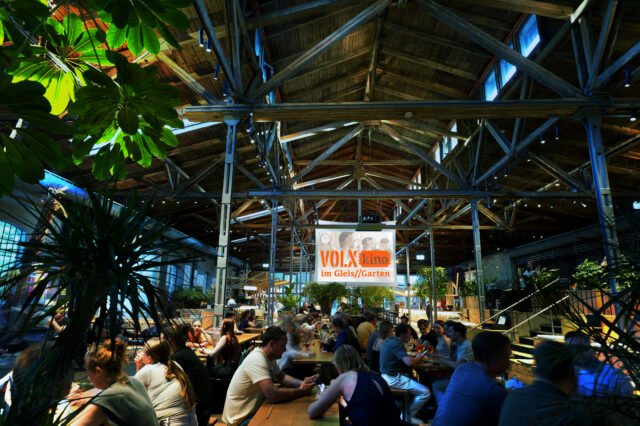 Innenaufnahme des VOLXkino im Gleis/Garten 2025: Menschen sitzen an langen Holztischen in einer großen, offenen Halle mit Holzdachkonstruktion, viel Grünpflanzen und gemütlicher Atmosphäre.