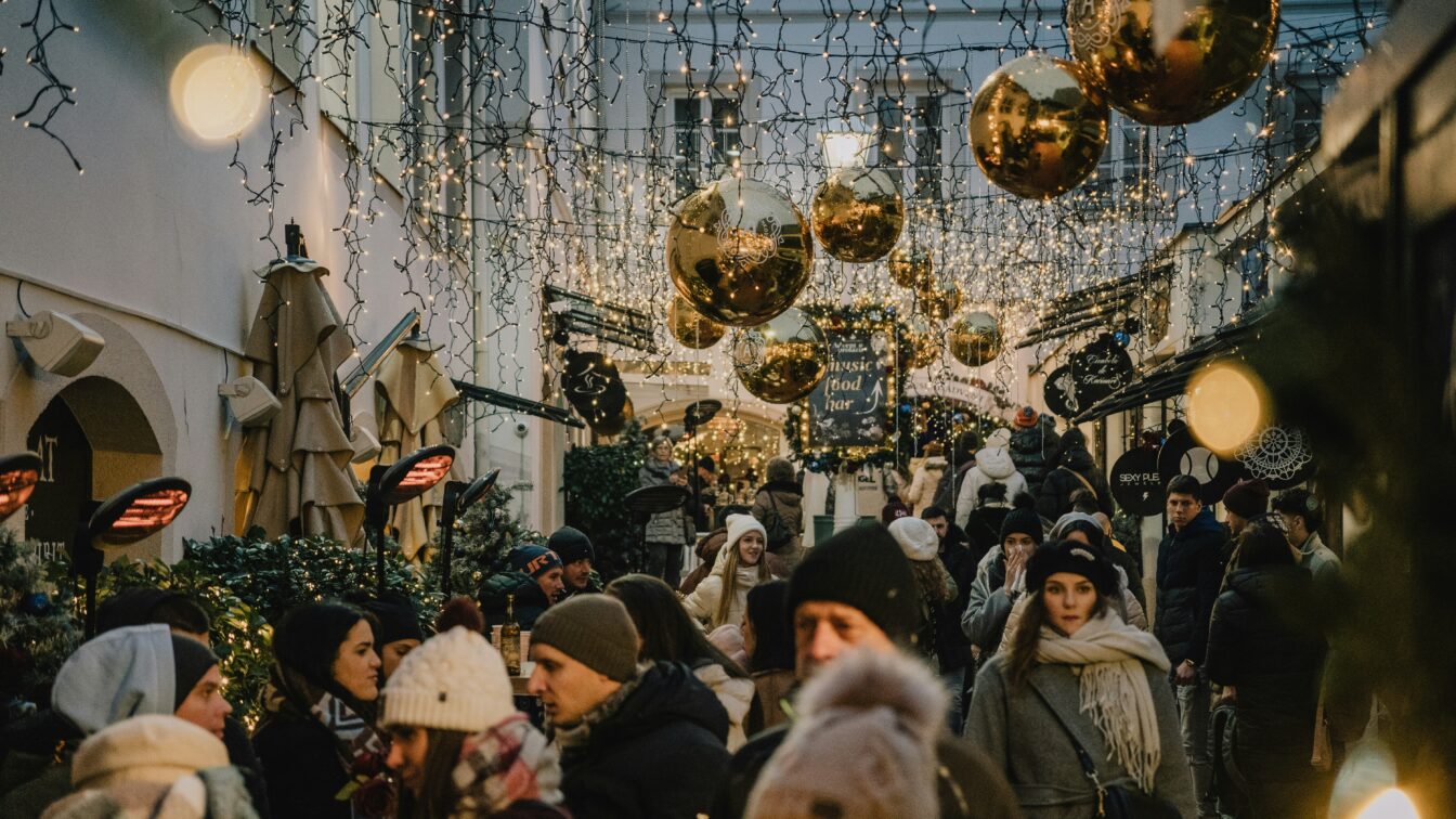 Belebter Weihnachtsmarkt in einer festlich geschmückten Gasse in Italien mit Lichterketten, goldenen Kugeln und Menschen in Winterkleidung.