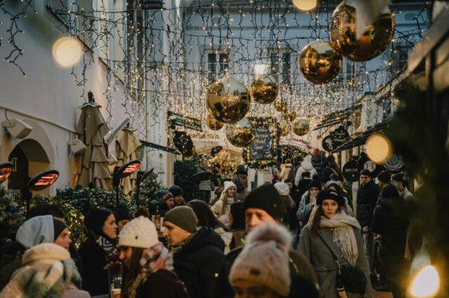 Belebter Weihnachtsmarkt in einer festlich geschmückten Gasse in Italien mit Lichterketten, goldenen Kugeln und Menschen in Winterkleidung.