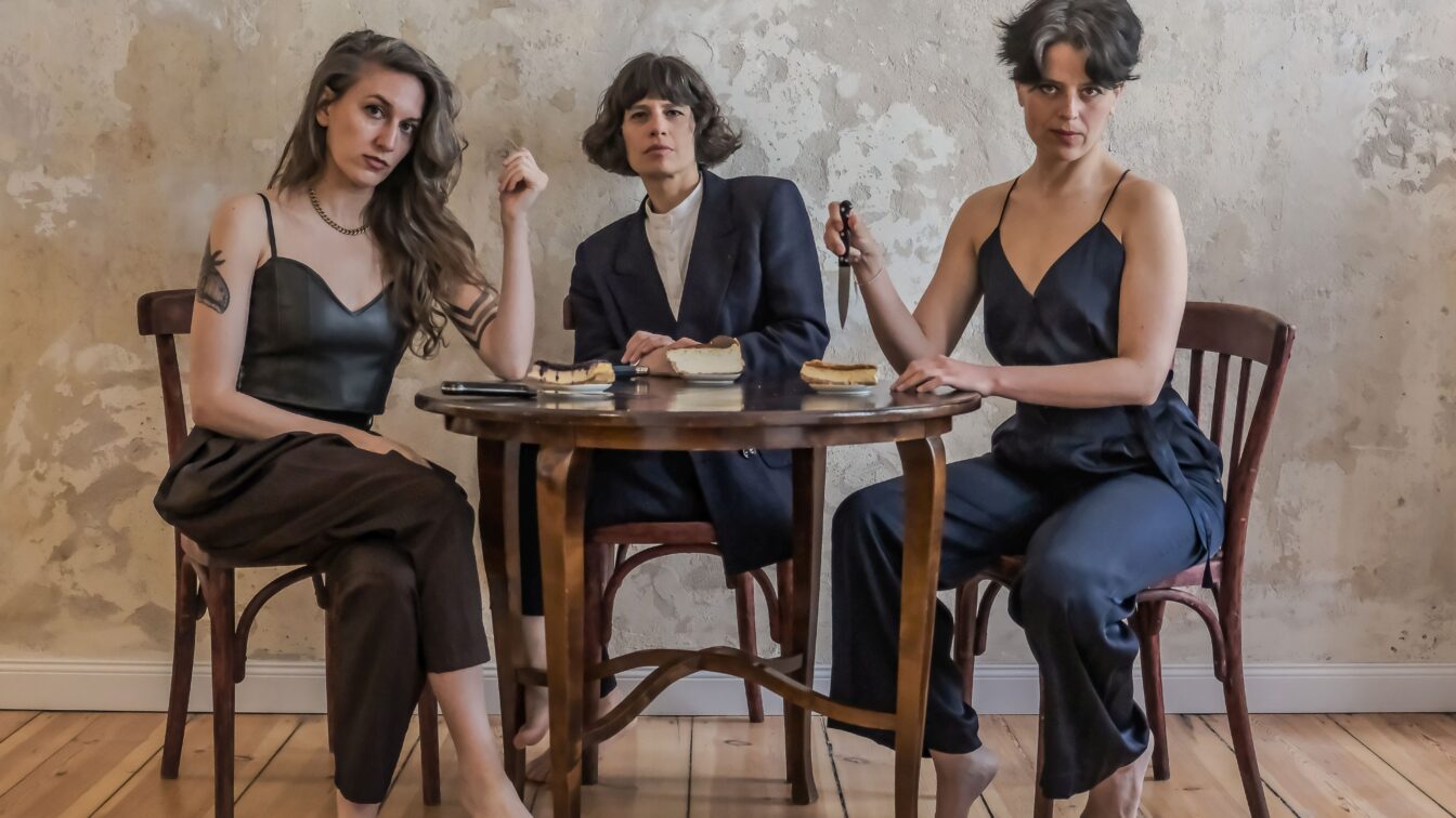 Drei Personen sitzen barfuß um einen kleinen Holztisch in einem schlichten Raum mit abgenutzter Wand. Sie blicken ernst in die Kamera. Auf dem Tisch liegen Teller mit Kuchenstücken und ein Messer, das eine der Personen in der Hand hält. Die Szene wirkt inszeniert und theatralisch, mit einem Hauch von Spannung