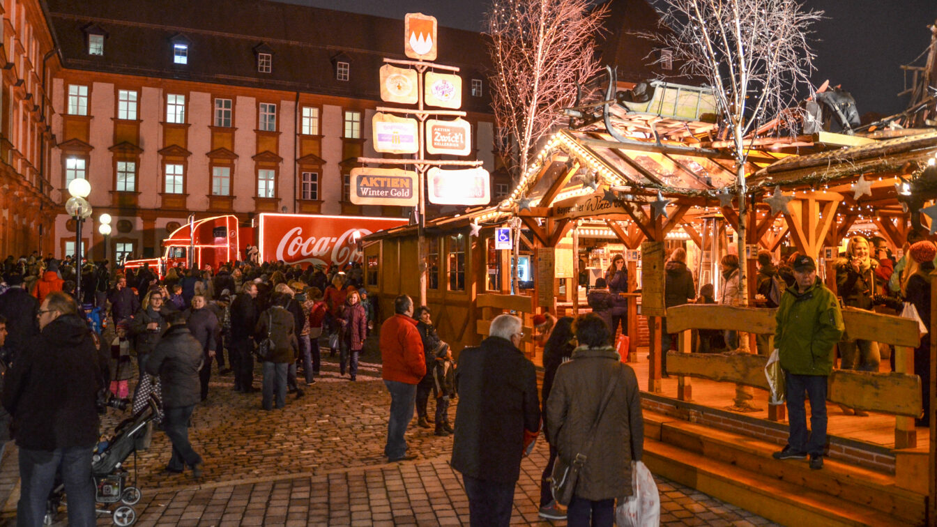 Menschen genießen einen winterlichen Abend im Bayreuther Winterdorf, mit festlich beleuchteten Holzhütten, geschmückten Bäumen und einem leuchtenden Coca-Cola-Weihnachtstruck im Hintergrund.